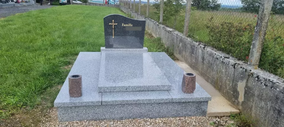 Monument funéraire en granit gris clair à stèle galbée et croix dorée, en bordure de cimetière
